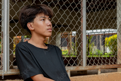 Εκτύπωση καμβά Asian teenboy in a black shirt sits with his knees pressed against a metal fence panel in a juvenile detention facility, awaiting further release, freedom and detention of people concept