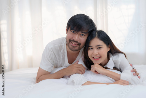 Young asian couple is lying on a bed with white sheets, smiling and enjoying their time together