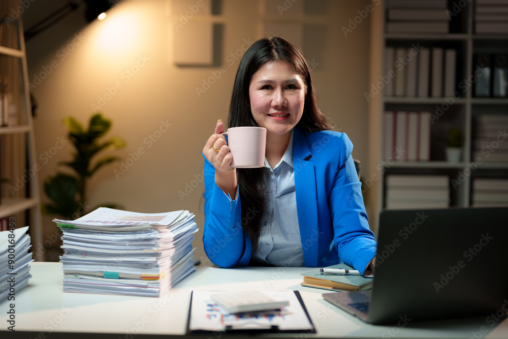 Asian businesswoman holding a coffee cup, relaxing, working on a laptop computer at the office in the evening. Overtime concept. Asian businesswoman holding a coffee cup, relaxing, working on a laptop computer at the office in the evening. Overtime concept.