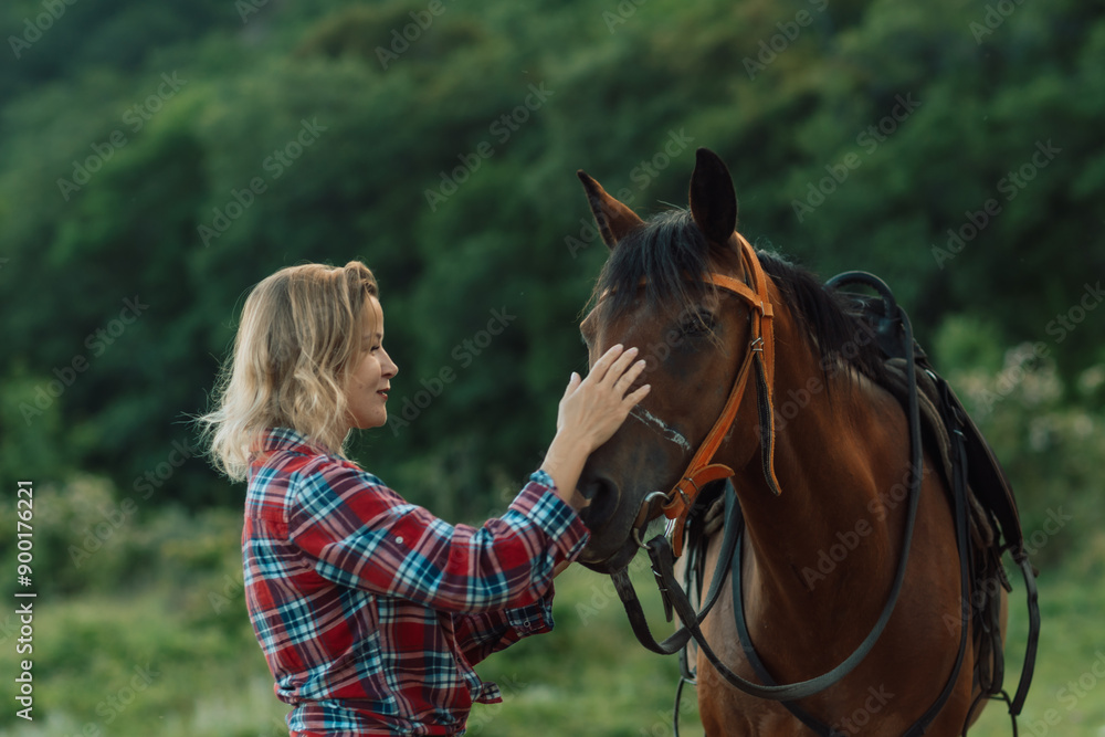 Obraz premium Happy blonde with horse in forest. Woman and a horse walking through the field during the day. Dressed in a plaid shirt and black leggings.