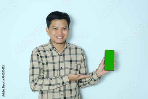 Young Asian man smiles happily while showing the green screen of his cell phone with his open right hand. Wearing a cream flannel shirt photo