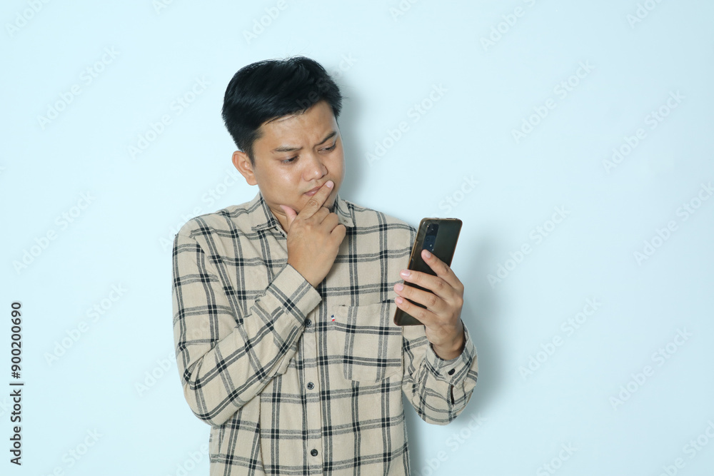 Young Asian man showing confused expression while looking to his mobile phone. Wearing a cream flannel shirt Young Asian man showing confused expression while looking to his mobile phone. Wearing a cream flannel shirt