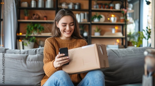 A smiling young woman is sitting on the sofa at home, holding a mobile phone and an open cardboard box in her hand, she is happy about the received package and the purchase.