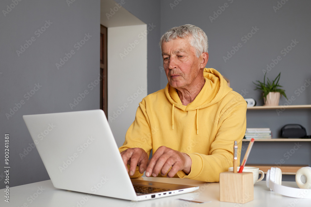 Senior mature Caucasian man wearing yellow hoodie typing on laptop working online doing his remote work retired male having freelance project at home Senior mature Caucasian man wearing yellow hoodie typing on laptop working online doing his remote work retired male having freelance project at home
