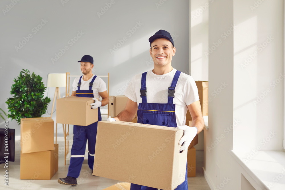 Workers from home removal service moving things to new house or apartment. Happy man in uniform overalls and cap carrying packed up cardboard box, looking at camera and smiling Workers from home removal service moving things to new house or apartment. Happy man in uniform overalls and cap carrying packed up cardboard box, looking at camera and smiling