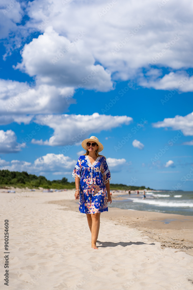 Summer vacation. Beautiful smiling woman wearing colorful dress and straw hat walking on sandy beach on sunny day. Summer walk among clouds. Front view Summer vacation. Beautiful smiling woman wearing colorful dress and straw hat walking on sandy beach on sunny day. Summer walk among clouds. Front view