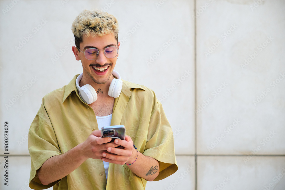 A man with a moustache and glasses is smiling while looking at his cell phone. He is wearing headphones and a yellow shirt