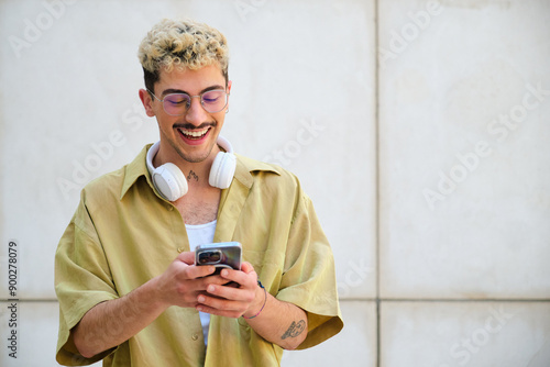 Obraz na płótnie A man with a moustache and glasses is smiling while looking at his cell phone. He is wearing headphones and a yellow shirt w salonie