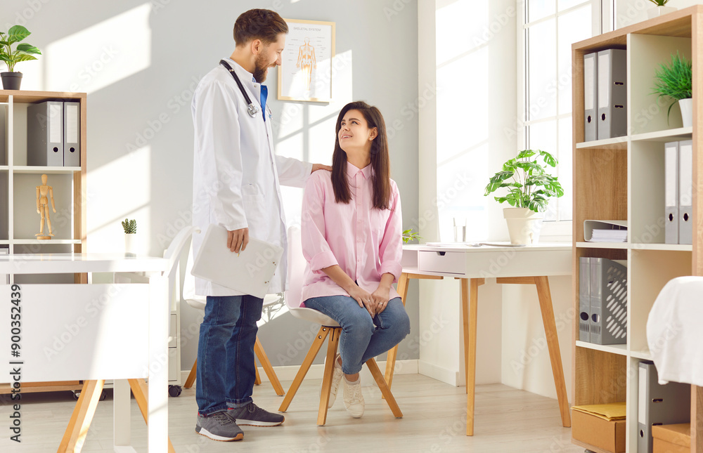 Female patient looks at her doctor with expression of trust and comfort during medical consultation. Doctor coat stands next to sitting patient and comforts her by putting his hand on her shoulder. Female patient looks at her doctor with expression of trust and comfort during medical consultation. Doctor coat stands next to sitting patient and comforts her by putting his hand on her shoulder.