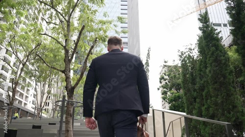 Back view of caucasian manager with formal suit walking up stairs while holding his suitcase in green city. Professional business man going to workiplace while climb stair in eco urban city. Urbane.. photo