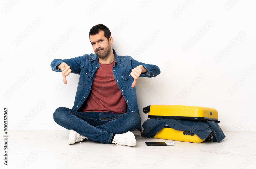 Caucasian handsome man with a suitcase full of clothes sitting on the floor at indoors showing thumb down with two hands Caucasian handsome man with a suitcase full of clothes sitting on the floor at indoors showing thumb down with two hands