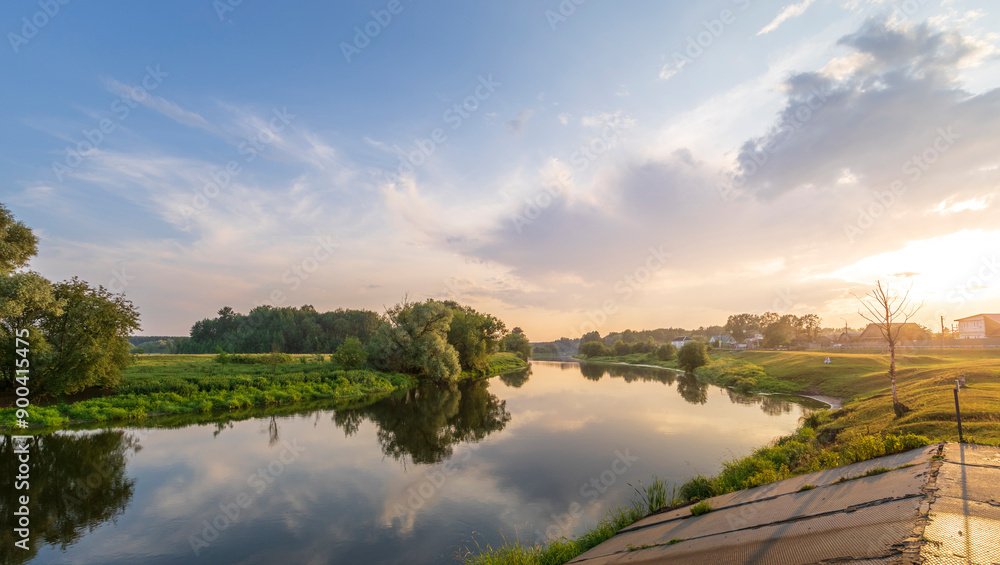 A serene rural river scene with lush trees by its banks. The sky is tinted orange and pink by the setting sun, casting a warm glow on the water A serene rural river scene with lush trees by its banks. The sky is tinted orange and pink by the setting sun, casting a warm glow on the water