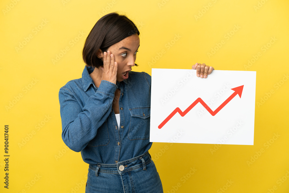 Young mixed race woman isolated on yellow background holding a sign with a growing statistics arrow symbol with surprised expression Young mixed race woman isolated on yellow background holding a sign with a growing statistics arrow symbol with surprised expression