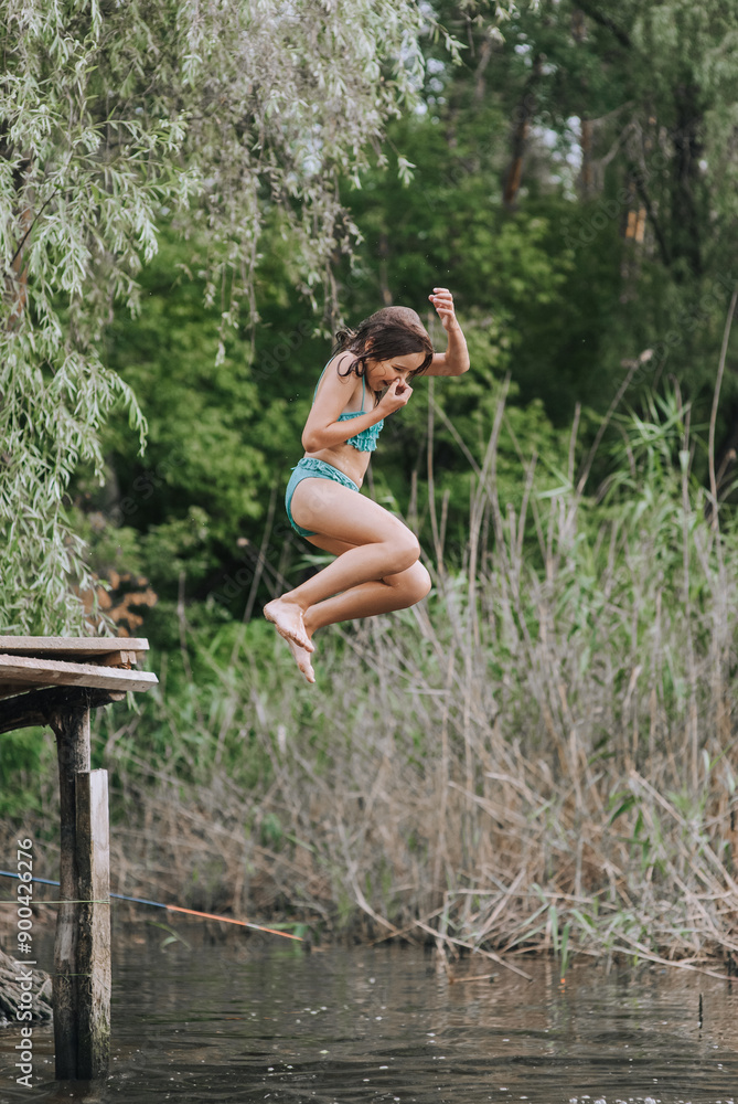 Fototapeta premium Athletic beautiful teenage girl, child jumping from a wooden bridge, pier into the water on a lake outdoors in nature in the summer on vacation. Photography, portrait, childhood concept, sport.