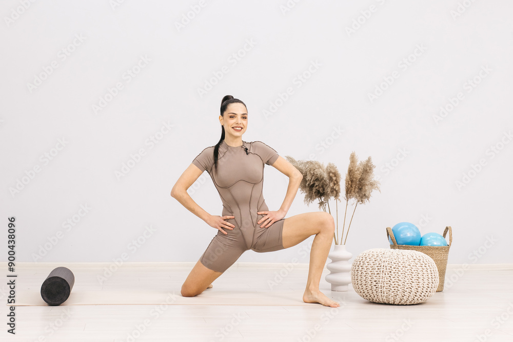 Fototapeta premium An athletic woman in sportswear is doing a workout to stretch her body and develop her muscles. Active girl doing fitness or yoga in home gym. Healthy body and sportswear concept.