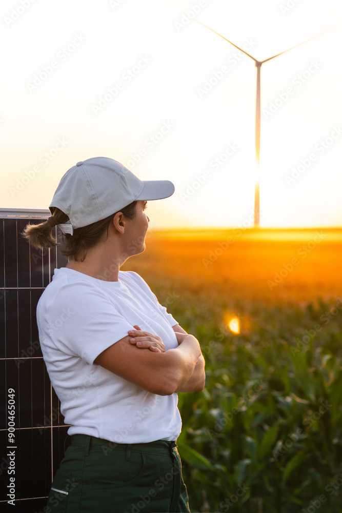 Fototapeta premium Woman wearing white cap stands next to solar panel at sunset. Wind turbines in the background