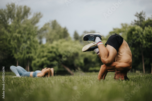 Obraz na plátně A man practices a headstand exercise in an open grassy park with trees and a woman exercising in the background on a sunny day