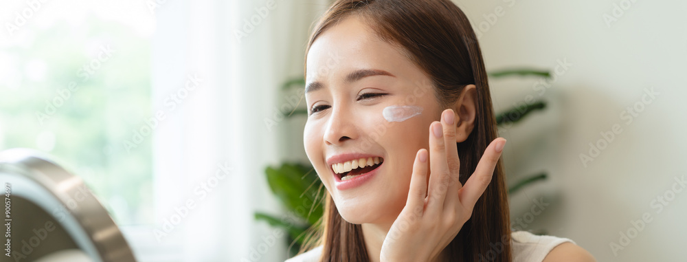 Facial beauty skin health care, smile of cute asian young woman looking at mirror, hand applying moisturizer lotion on her face, female putting cream treatment before makeup cosmetic routine at home. Facial beauty skin health care, smile of cute asian young woman looking at mirror, hand applying moisturizer lotion on her face, female putting cream treatment before makeup cosmetic routine at home.
