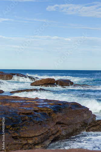 The image captures a serene coastal scene with rocky formations by the sea, gentle waves lapping against the shore, and a clear blue sky with scattered clouds in the background. photo