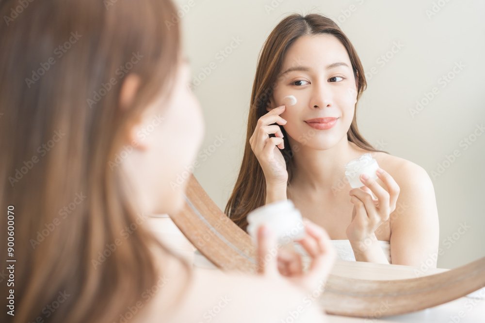 Facial beauty skin care, smile of pretty asian young woman in bathrobe looking at mirror, hand applying moisturizer lotion on her face, holding jar of skin cream before makeup cosmetic routine at home Facial beauty skin care, smile of pretty asian young woman in bathrobe looking at mirror, hand applying moisturizer lotion on her face, holding jar of skin cream before makeup cosmetic routine at home