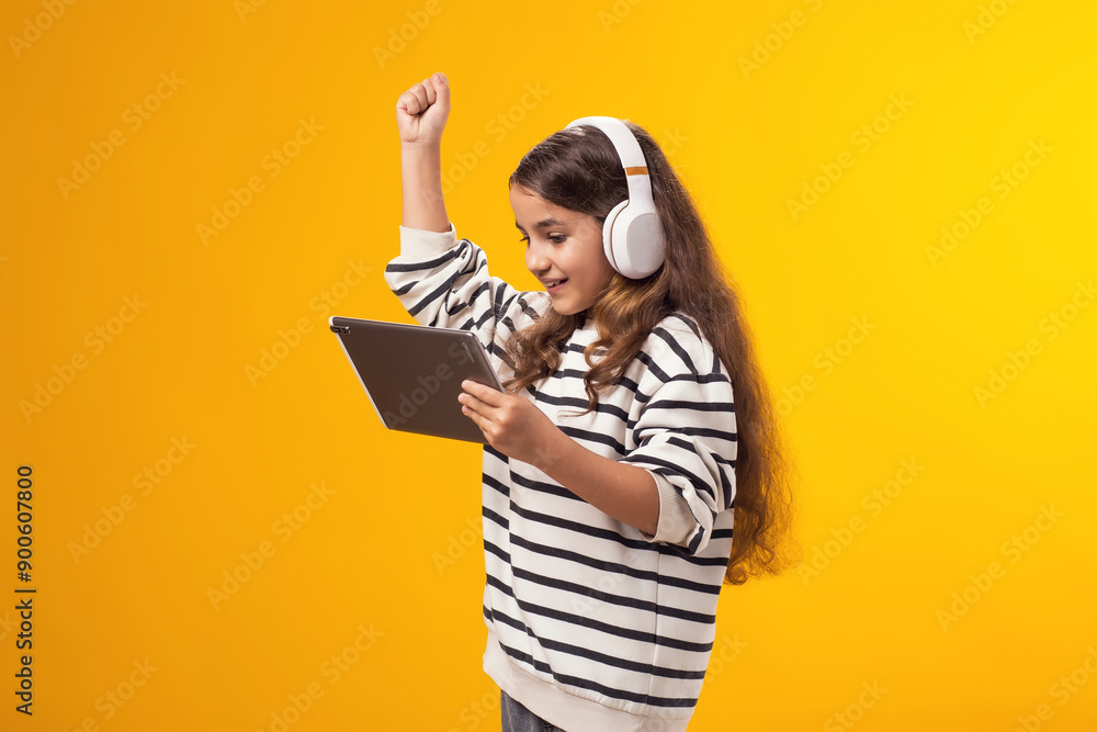 Happy girl with headphones holding tablet in hand and showing winner gesture on yellow background. Lifestyle, leasure and gadget addiction concept Happy girl with headphones holding tablet in hand and showing winner gesture on yellow background. Lifestyle, leasure and gadget addiction concept