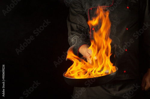 A chef cooking food on frying pan with fire flame or flambe. Concept of cooking food for lunch in pub. Space for recipe on black background