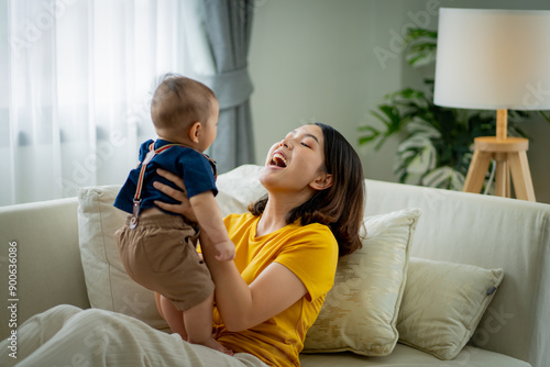 A woman is holding a baby and smiling. The baby is wearing a blue shirt. The woman is sitting on a couch