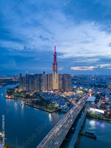 Landmark building illuminated night drone shot with Saigon river boat traffic and road traffic on the bridge below in vertical format. A dynamic welcome establishing Ho Chi Minh City, Vietnam.