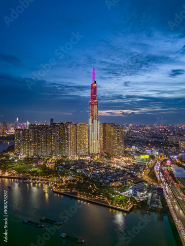 Landmark building illuminated night drone shot with Saigon river boat traffic and road traffic on the bridge below in vertical format. A dramatic welcome to Ho Chi Minh City, Vietnam.