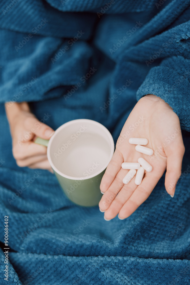 Person holding cup of pills and coffee wrapped in blanket with pillows Person holding cup of pills and coffee wrapped in blanket with pillows