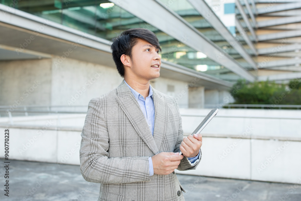 Asian businessman stands in front of a modern building, holding a tablet in his hands. He is looking up, seemingly in deep thought or listening to someone off-screen, copy space Asian businessman stands in front of a modern building, holding a tablet in his hands. He is looking up, seemingly in deep thought or listening to someone off-screen, copy space