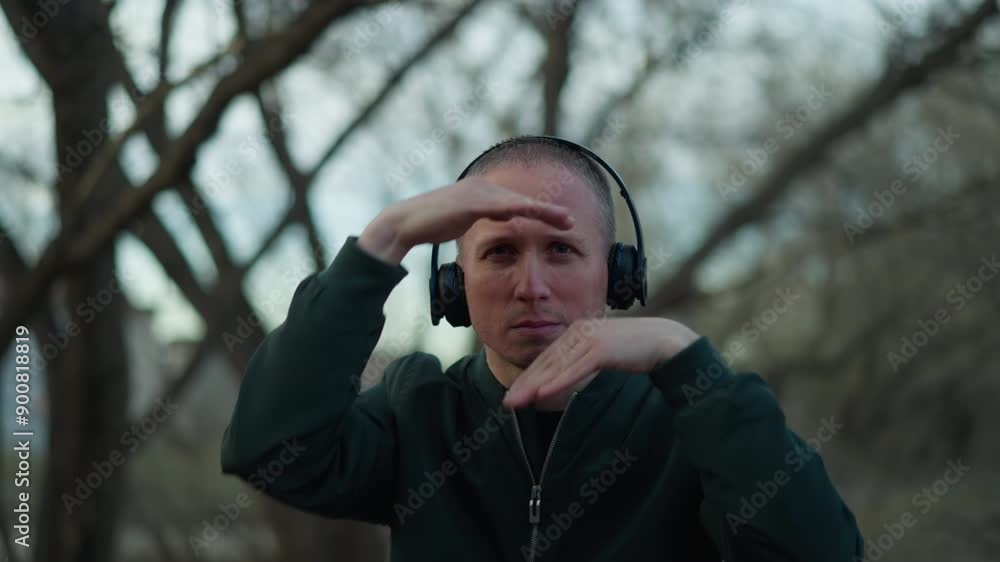 A man in a green jacket and headphones frames his face with his hands in a forest, his focused expression and distinctive pose stand out against the serene wooded background A man in a green jacket and headphones frames his face with his hands in a forest, his focused expression and distinctive pose stand out against the serene wooded background