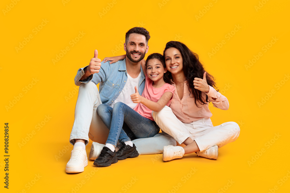 A happy family of three, a mother, father, and young daughter, sit on a yellow background, smiling and giving thumbs up. The family is dressed in casual clothes, they are all looking at the camera. A happy family of three, a mother, father, and young daughter, sit on a yellow background, smiling and giving thumbs up. The family is dressed in casual clothes, they are all looking at the camera.