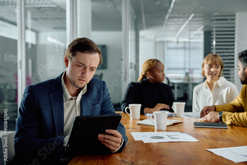 Four focused young multiracial adults in businesswear sitting in meeting using digital tablet at office