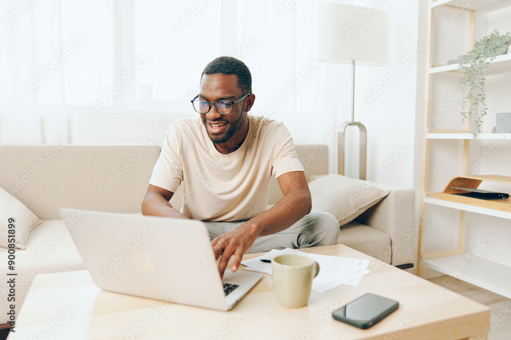 Smiling African American Man Working on Laptop from Home in Modern Living Room The young black freelancer is sitting on the sofa, typing on his computer, fully immersed in his online education His Smiling African American Man Working on Laptop from Home in Modern Living Room The young black freelancer is sitting on the sofa, typing on his computer, fully immersed in his online education His