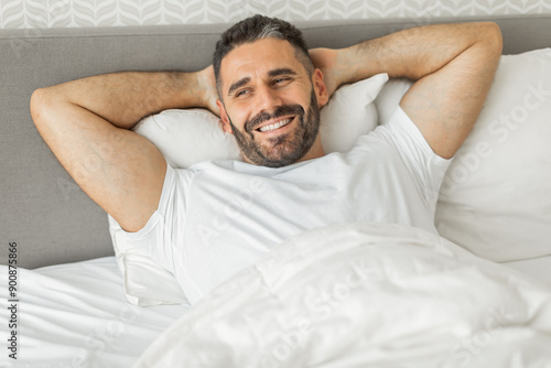 A man with a beard and short hair is lying in bed, smiling and looking directly at the camera. He is wearing a white t-shirt and his hands are behind his head, resting on a white pillow. © Prostock-studio