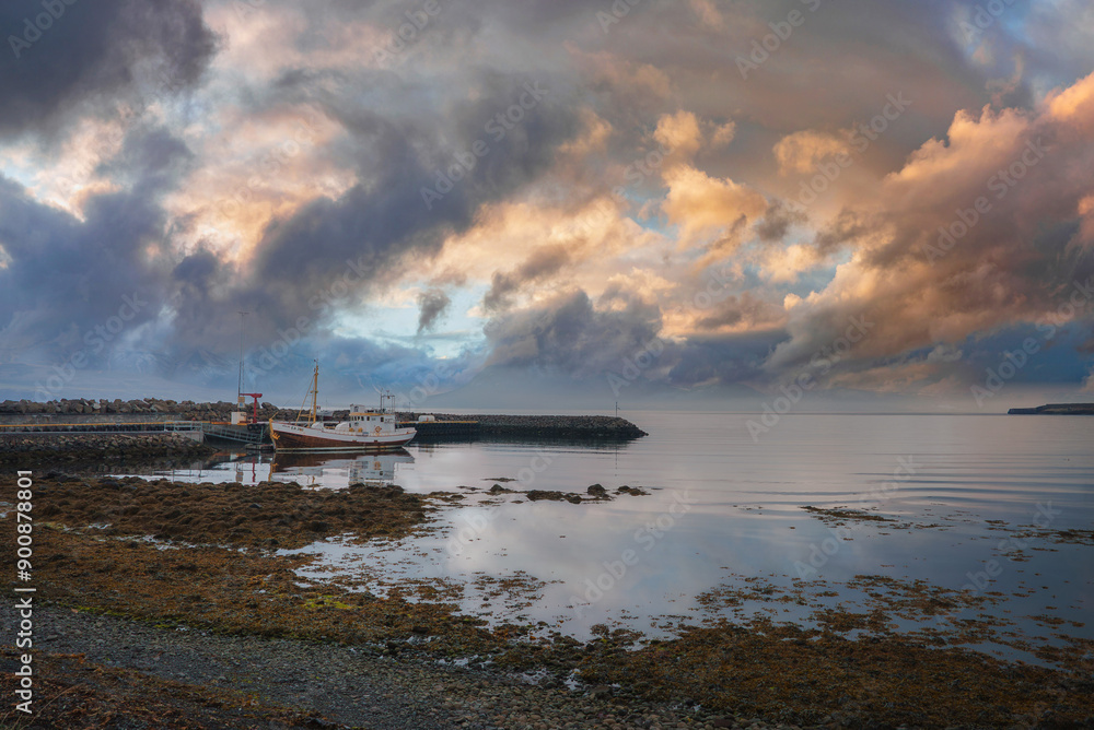 A tranquil harbor in Iceland with a docked fishing boat, colorful clouds reflecting on calm waters, and a shoreline covered in seaweed and rocks. A tranquil harbor in Iceland with a docked fishing boat, colorful clouds reflecting on calm waters, and a shoreline covered in seaweed and rocks.