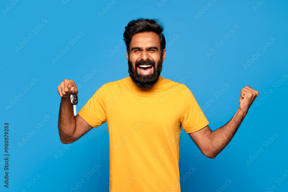Fototapeta premium Hindu man with a beard is smiling and holding up a car key in his right hand while his left fist is clenched in the air. He is wearing a yellow t-shirt and is standing in front of a blue background.