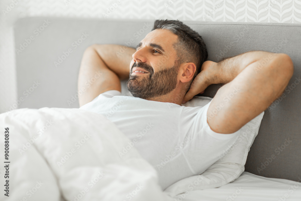 A man with a beard lies in bed with his hands behind his head, looking off to the side with a relaxed expression on his face. He is wearing a white t-shirt and is covered by a white duvet. A man with a beard lies in bed with his hands behind his head, looking off to the side with a relaxed expression on his face. He is wearing a white t-shirt and is covered by a white duvet.