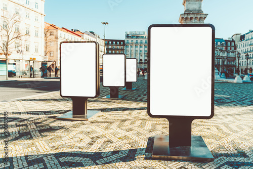 A row of blank billboards in a city square, ready for advertisement mockups. The billboards stand on a patterned pavement with surrounding historical buildings and a clear blue sky photo