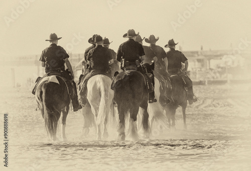 A Group of Police Officers on horseback on beach in late afternoon sun. The image is in black and white vintage tones
