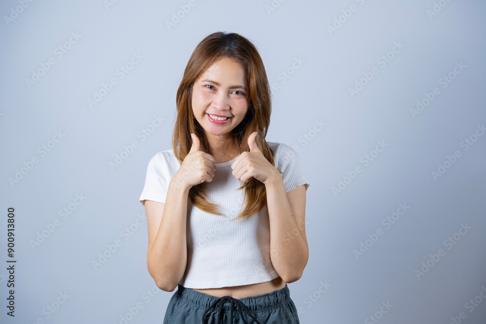 Smiling young woman giving two thumbs up gesture, wearing casual white shirt and grey pants against a white background. Smiling young woman giving two thumbs up gesture, wearing casual white shirt and grey pants against a white background.
