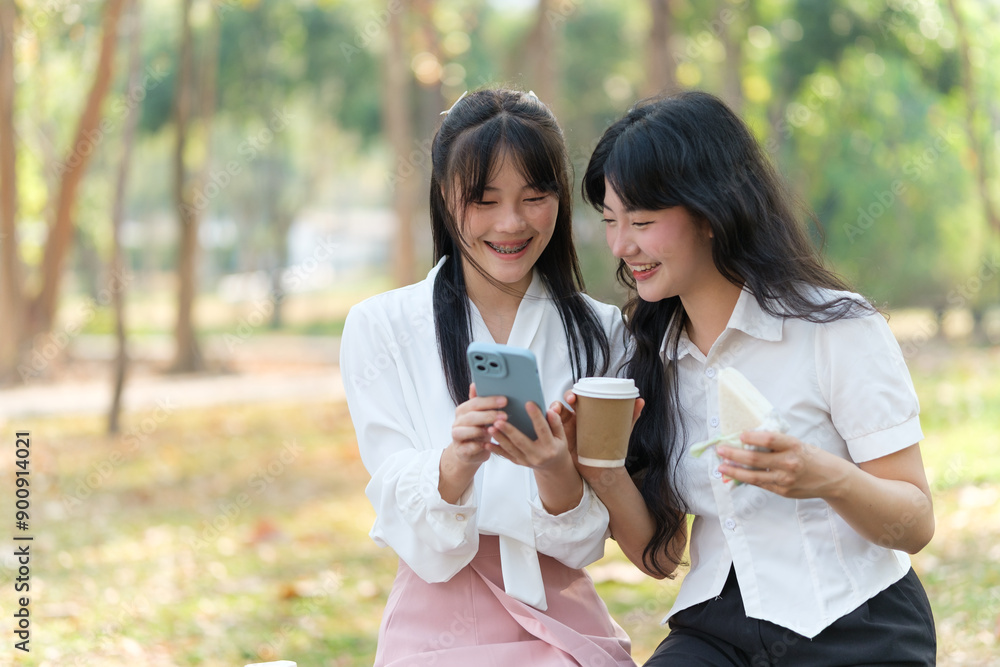 Two young women enjoy a casual coffee break in a park, sharing smiles as they look at a smartphone. Both hold coffee cups, with one holding a sandwich. Two young women enjoy a casual coffee break in a park, sharing smiles as they look at a smartphone. Both hold coffee cups, with one holding a sandwich.