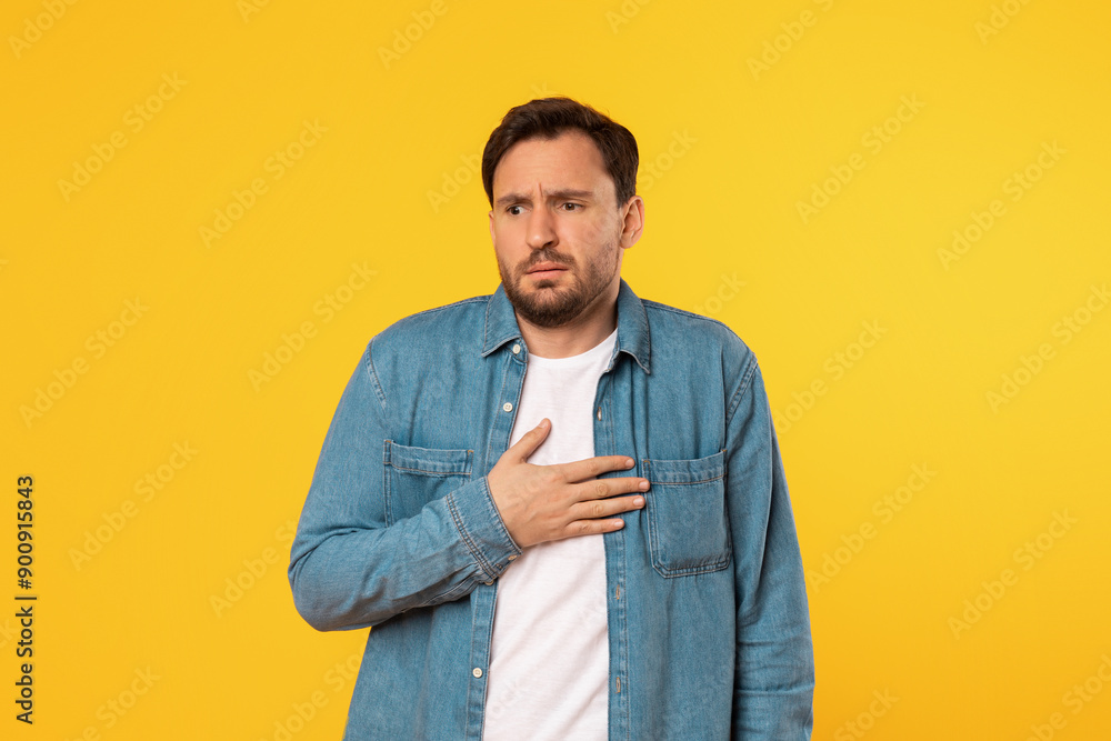 A man wearing a blue denim shirt and a white undershirt stands in front of a bright yellow background. He has a concerned expression on his face and is holding his hand to his chest. A man wearing a blue denim shirt and a white undershirt stands in front of a bright yellow background. He has a concerned expression on his face and is holding his hand to his chest.