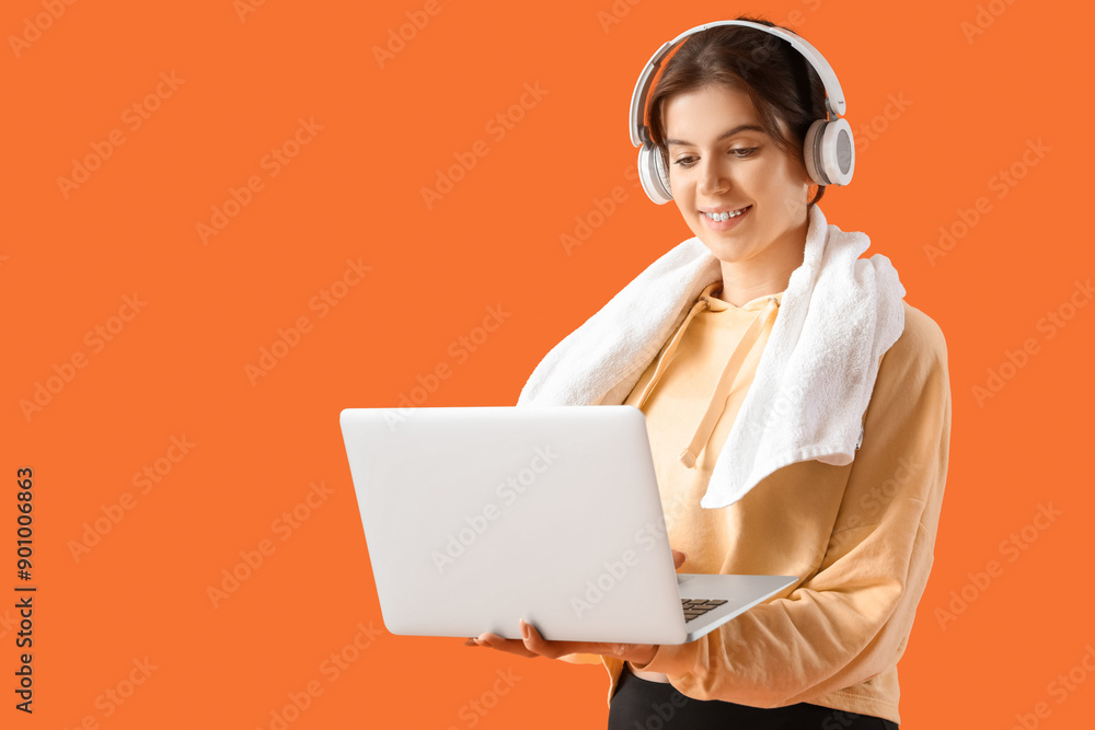 Sporty young woman in headphones with towel using laptop on orange background Sporty young woman in headphones with towel using laptop on orange background