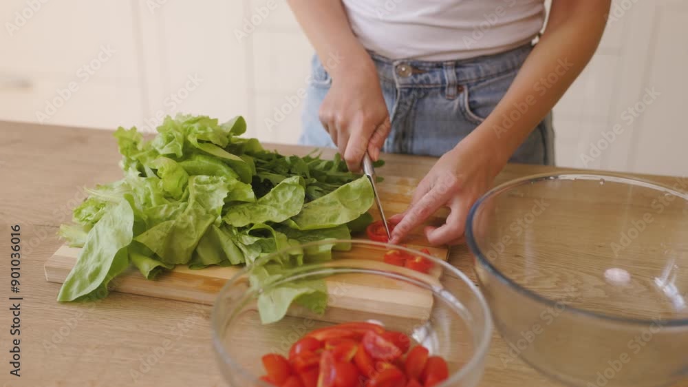 Woman is preparing a salad in the kitchen. The close-up shot captures their hands as they slice a cherry tomato on a cutting board next to a pile of fresh greens, such as lettuce and arugula. Woman is preparing a salad in the kitchen. The close-up shot captures their hands as they slice a cherry tomato on a cutting board next to a pile of fresh greens, such as lettuce and arugula.