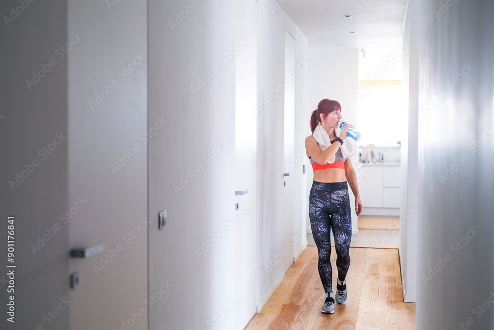 Beautiful redhead woman drinking water, resting after home fitness routine. Home workout on gym mt in living room, sporty woman wearing sportswear. Beautiful redhead woman drinking water, resting after home fitness routine. Home workout on gym mt in living room, sporty woman wearing sportswear.