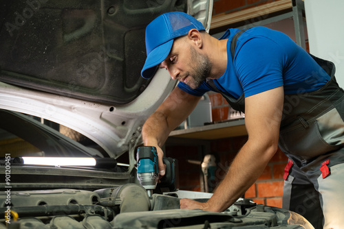 A mechanic works on a car engine in a garage. He is using a power tool to tighten bolts and screws. The mechanic is wearing work overalls and a blue baseball cap