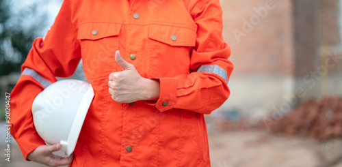 Fotografía Action of a construction foreman in orange coverall uniform is giving thumb up and holding white safety helmet, standing at construction site