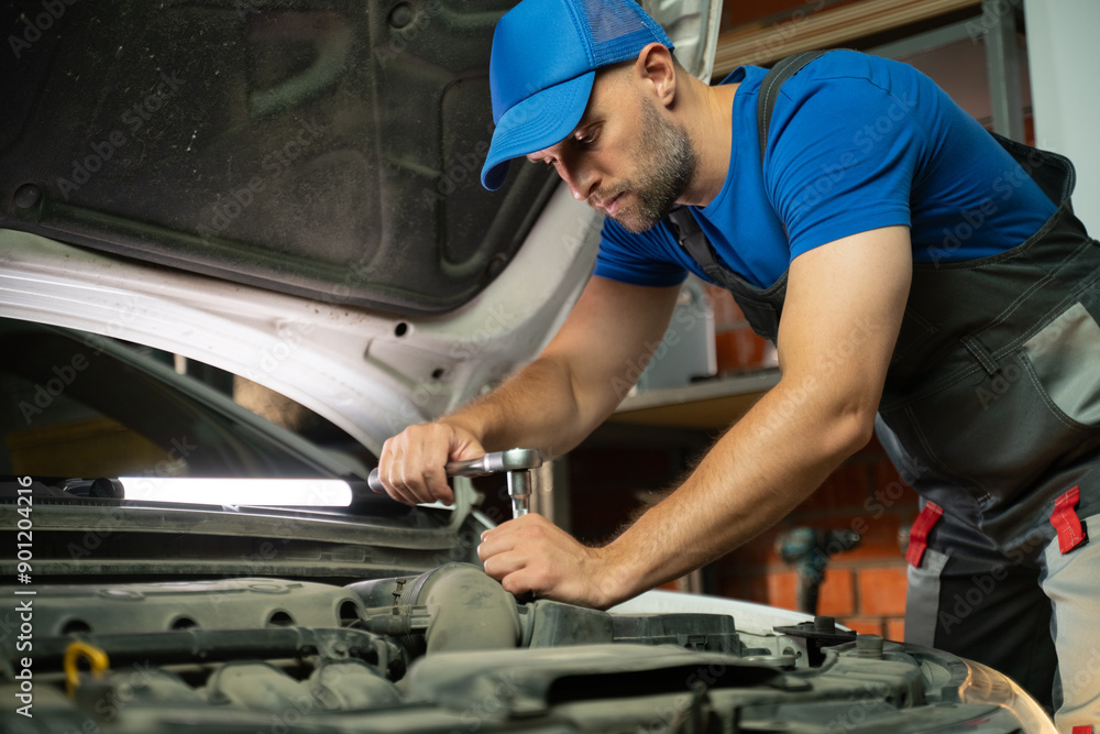 The mechanic works with the car engine in the garage. The mechanic uses a wrench to tighten the bolt. The hood of the car is open and the engine is illuminated by a bright light. The mechanic has a The mechanic works with the car engine in the garage. The mechanic uses a wrench to tighten the bolt. The hood of the car is open and the engine is illuminated by a bright light. The mechanic has a